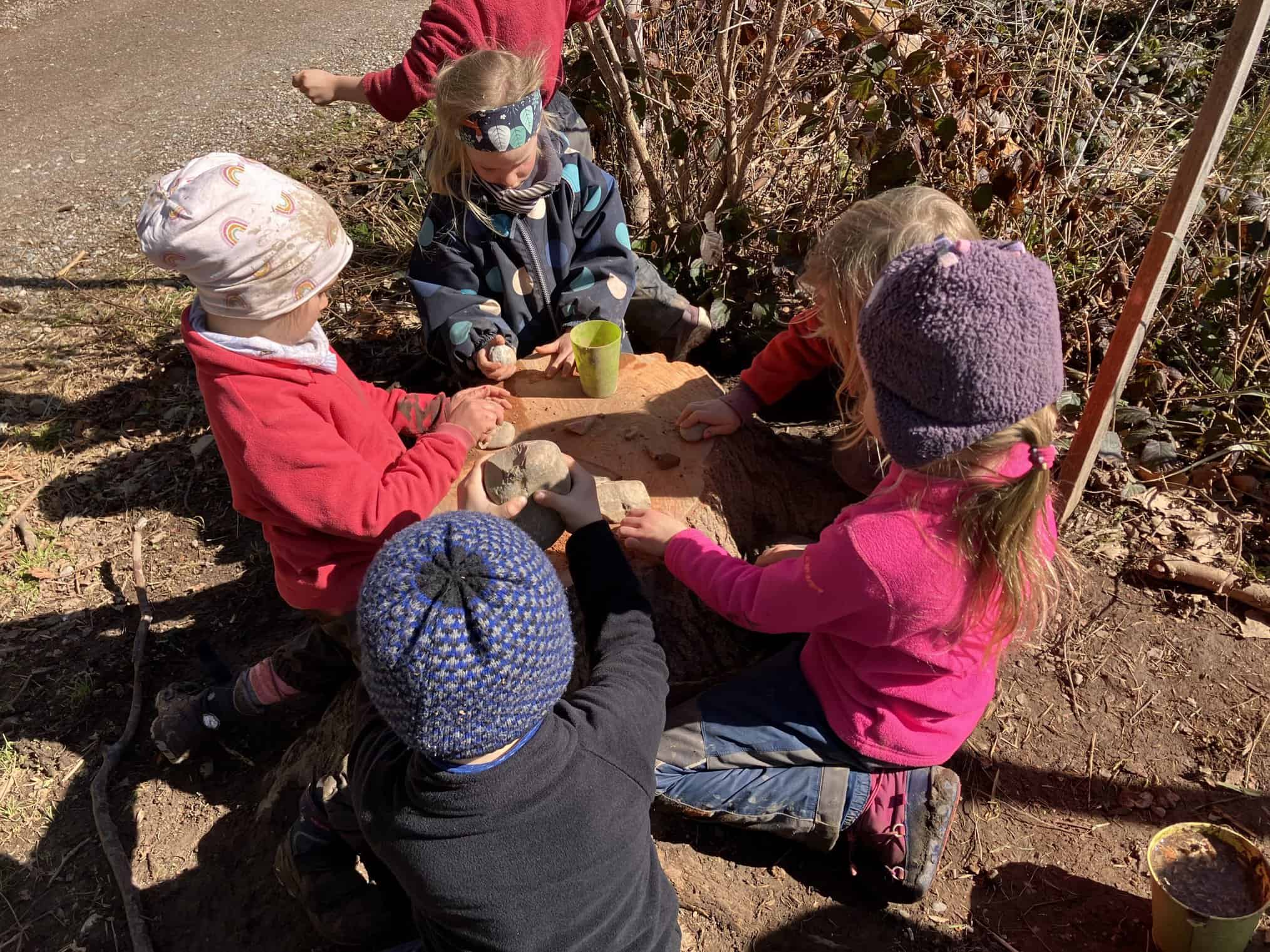 Kinder sitzen um einen abgesägten Holzstumpf und spielen auf diesem.