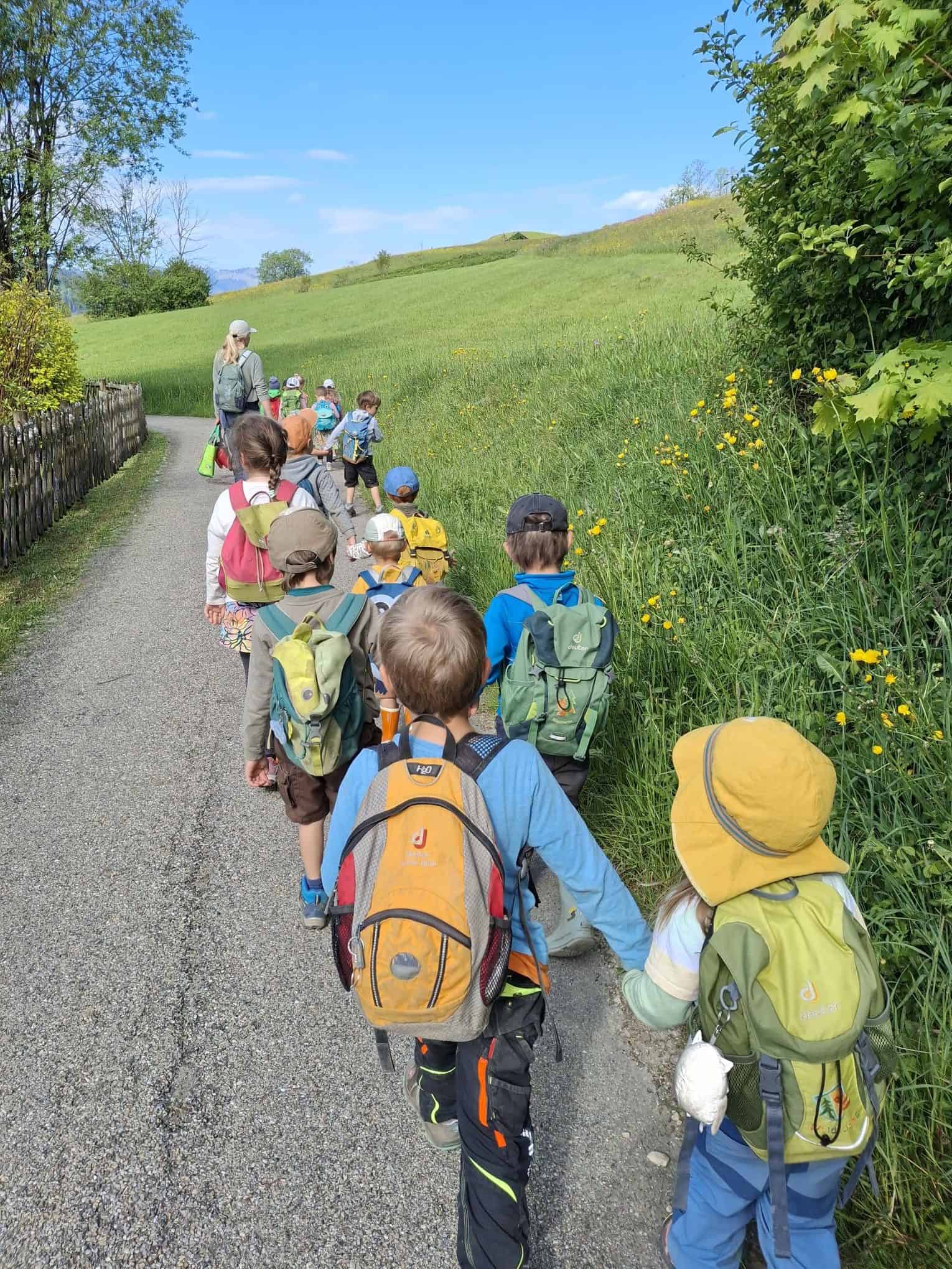 Kindergartenkinder laufen in Zweierreihen mit Rucksäcken auf einem Weg neben einem grünen Feld.