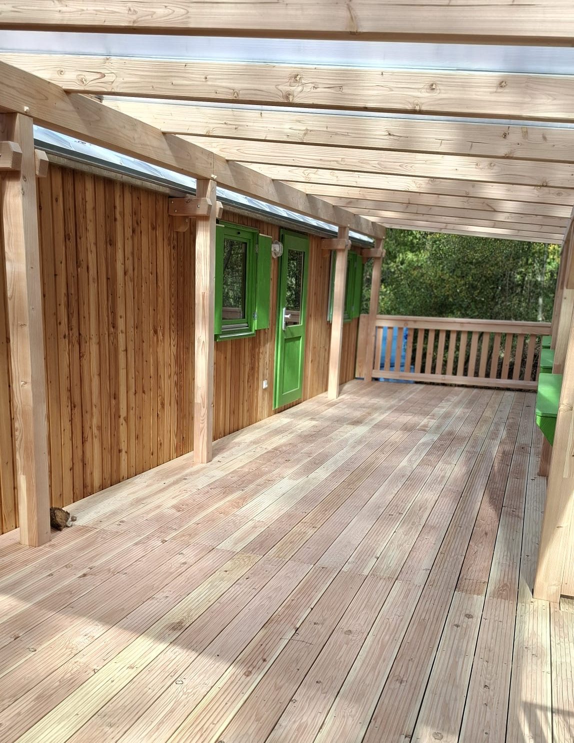 Ein hellbrauner Bauwagen aus Holz mit grünen Türen und Fenstern und einer großen Terrasse davor.