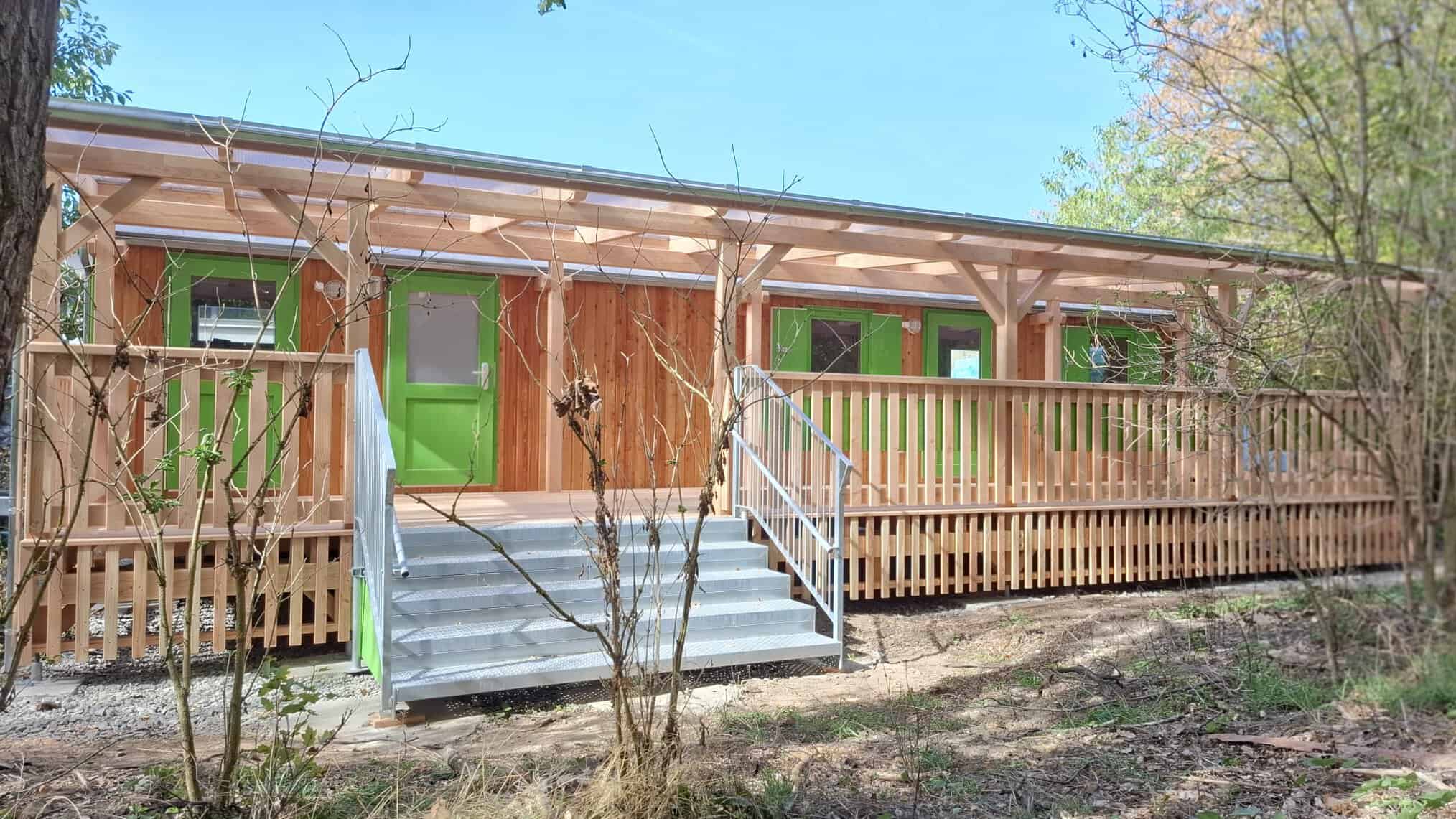 Ein hellbrauner Bauwagen aus Holz mit grünen Türen und Fenstern und einer großen Terrasse davor.